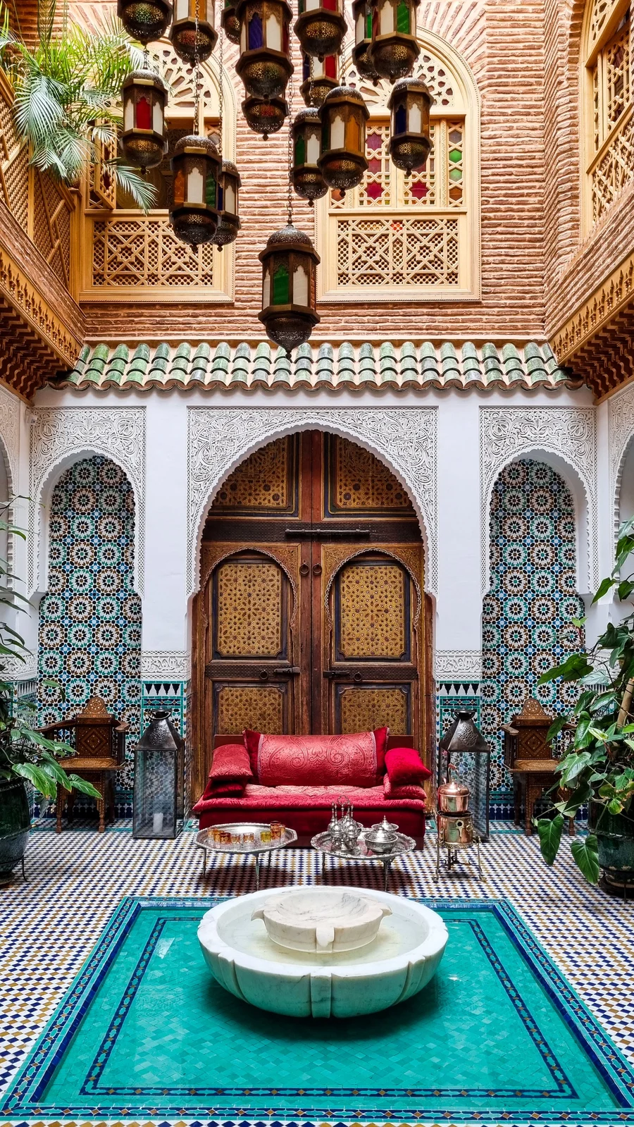 A traditional Moroccan riad courtyard in Marrakesh, with zellige tilework, an arched wooden door, and a central marble fountain