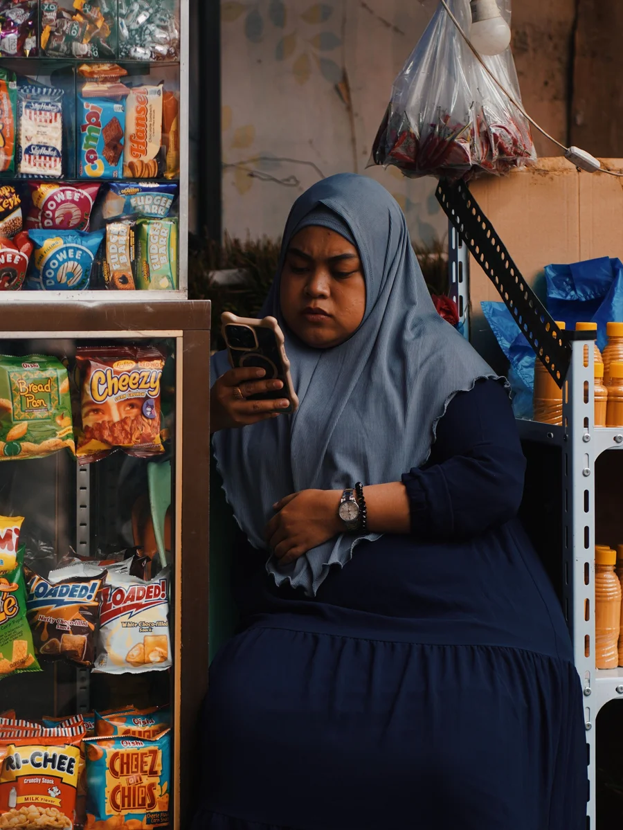 A shop owner in a hijab checking her phone at the counter of a small, modest neighborhood shop, shelves of everyday goods around her